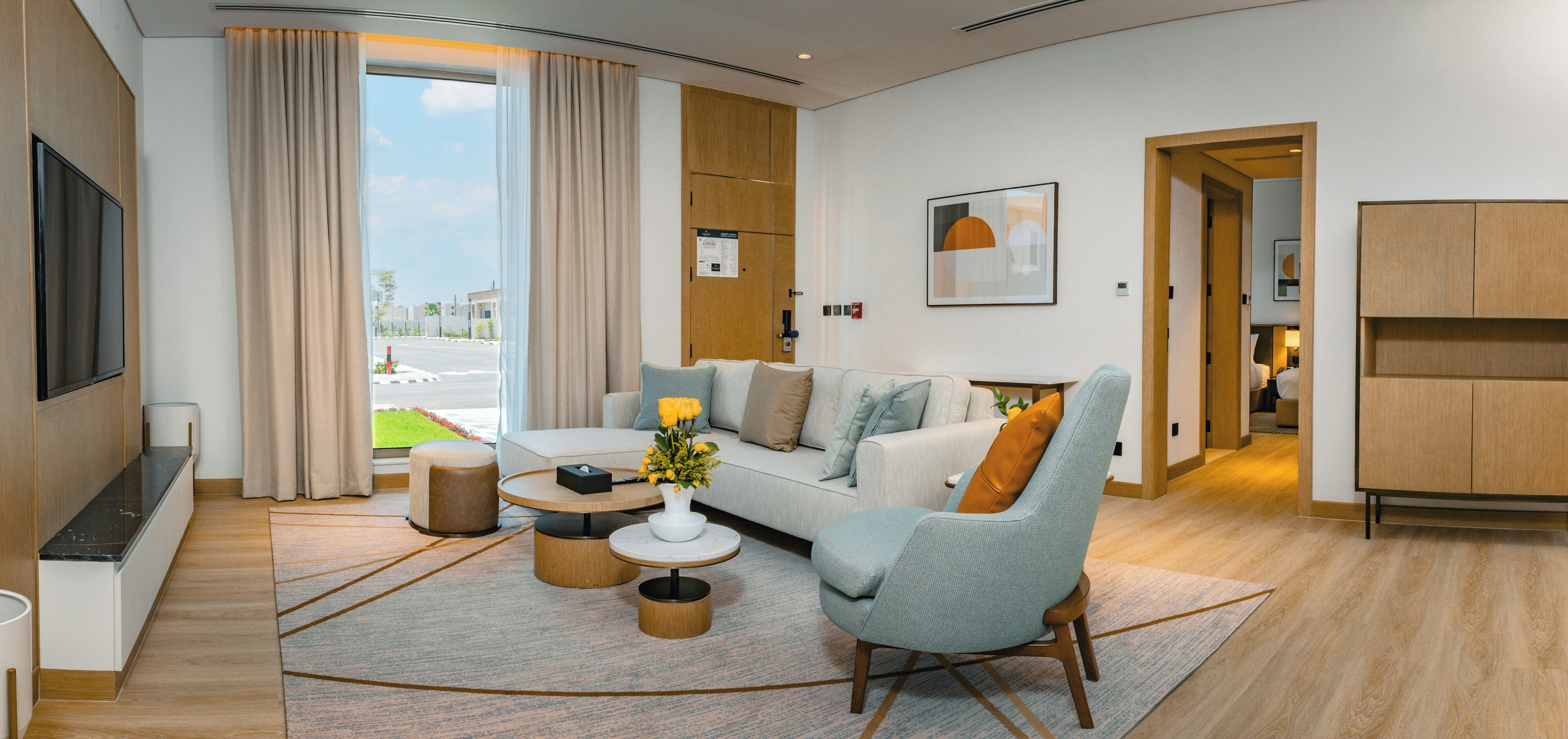 A modern living room with a sofa, armchairs, a coffee table, and a TV. There is a large window with curtains, a wooden cabinet, and a small table with a vase of flowers.