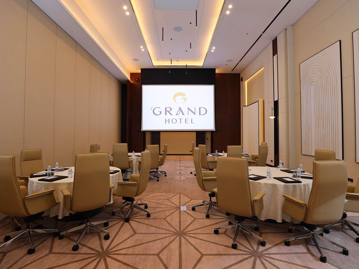 Een vergaderruimte met meerdere tafels en stoelen, een groot scherm waarop het logo van 'Grand Hotel' staat, en een vloerkleed met een geometrisch patroon.