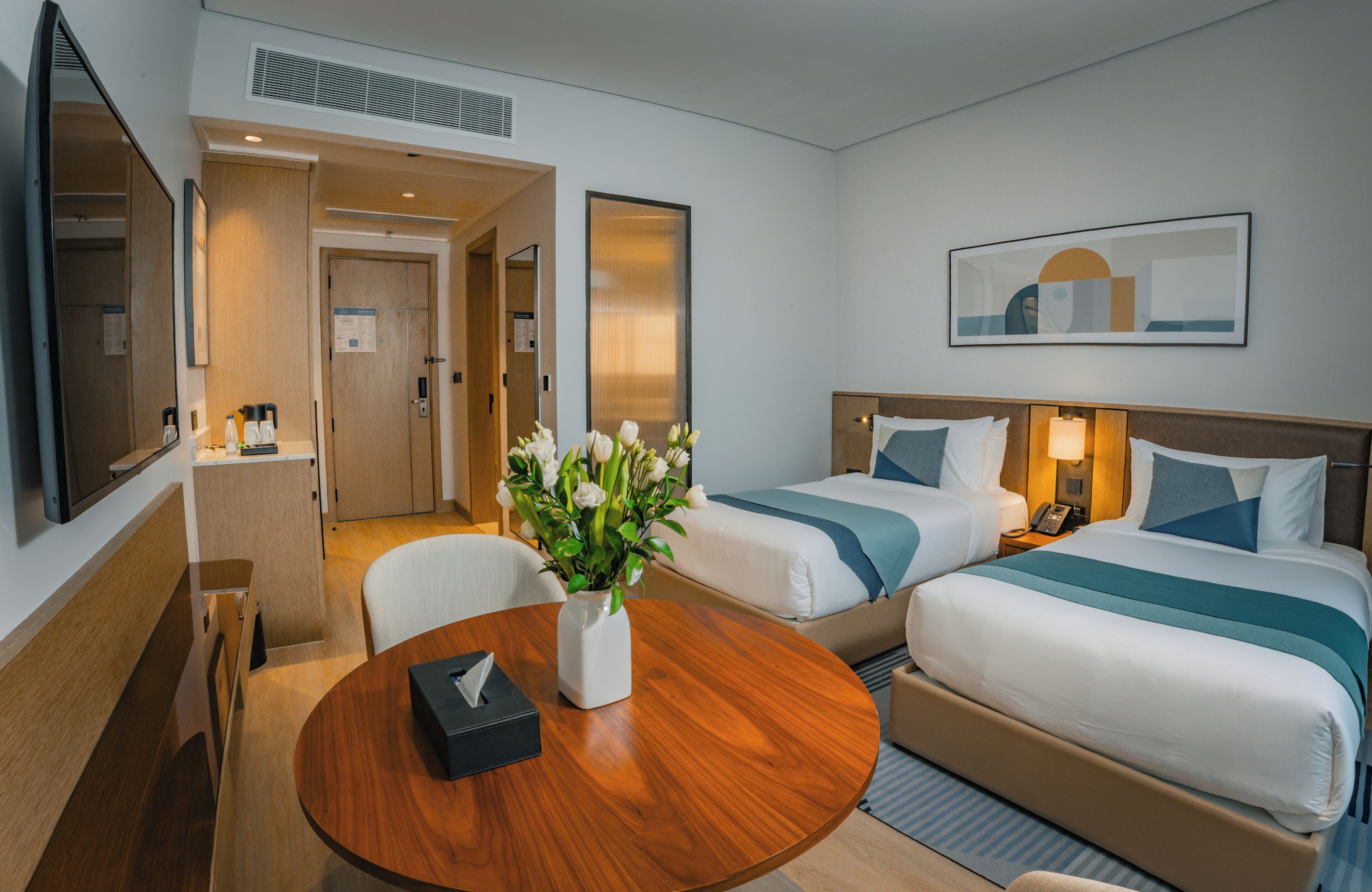 A hotel room with two beds, a wooden table with a vase of flowers, a desk with a telephone, and a mirror.
