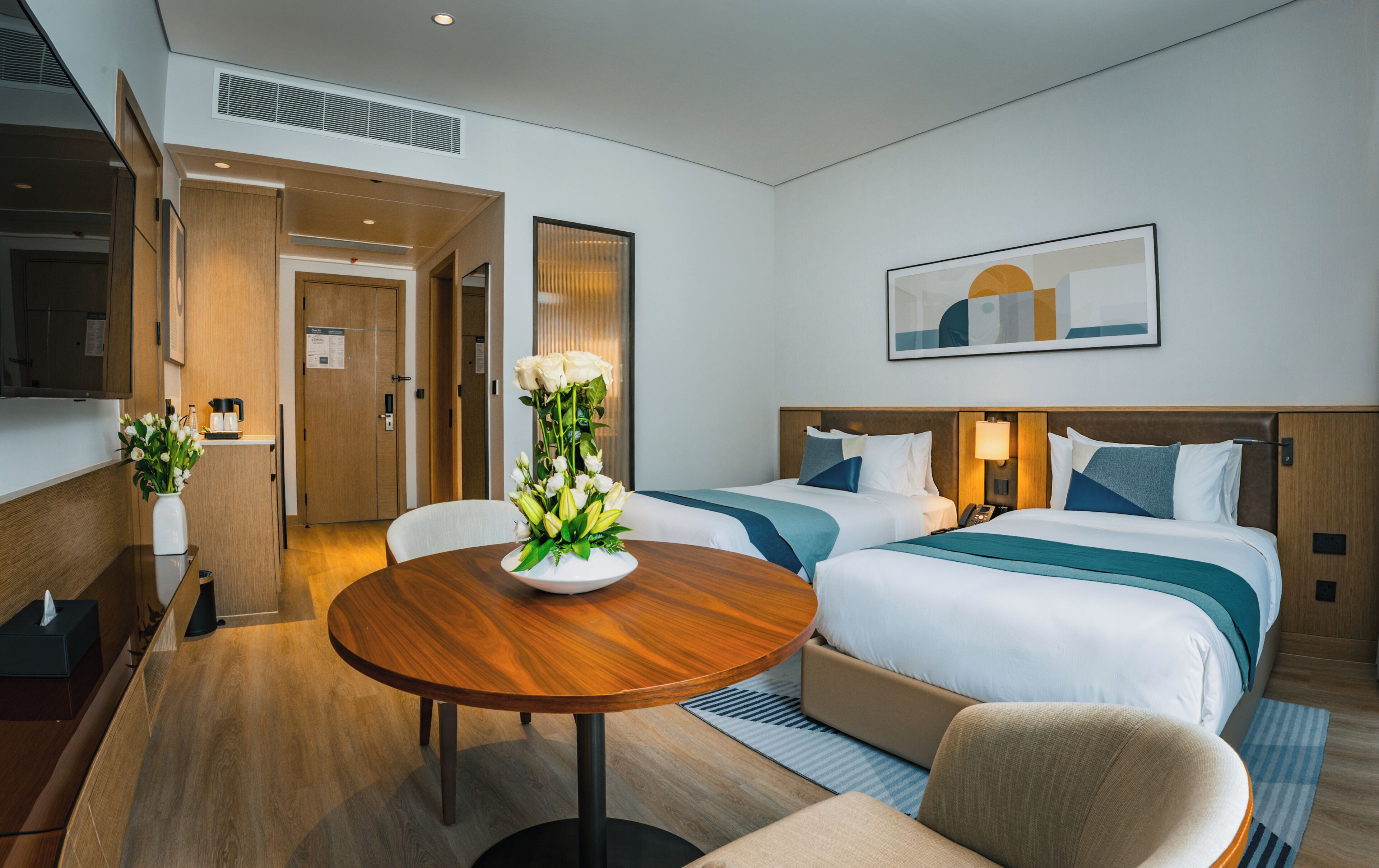 A hotel room with two beds, a dining table, a chair, and a vase of flowers.