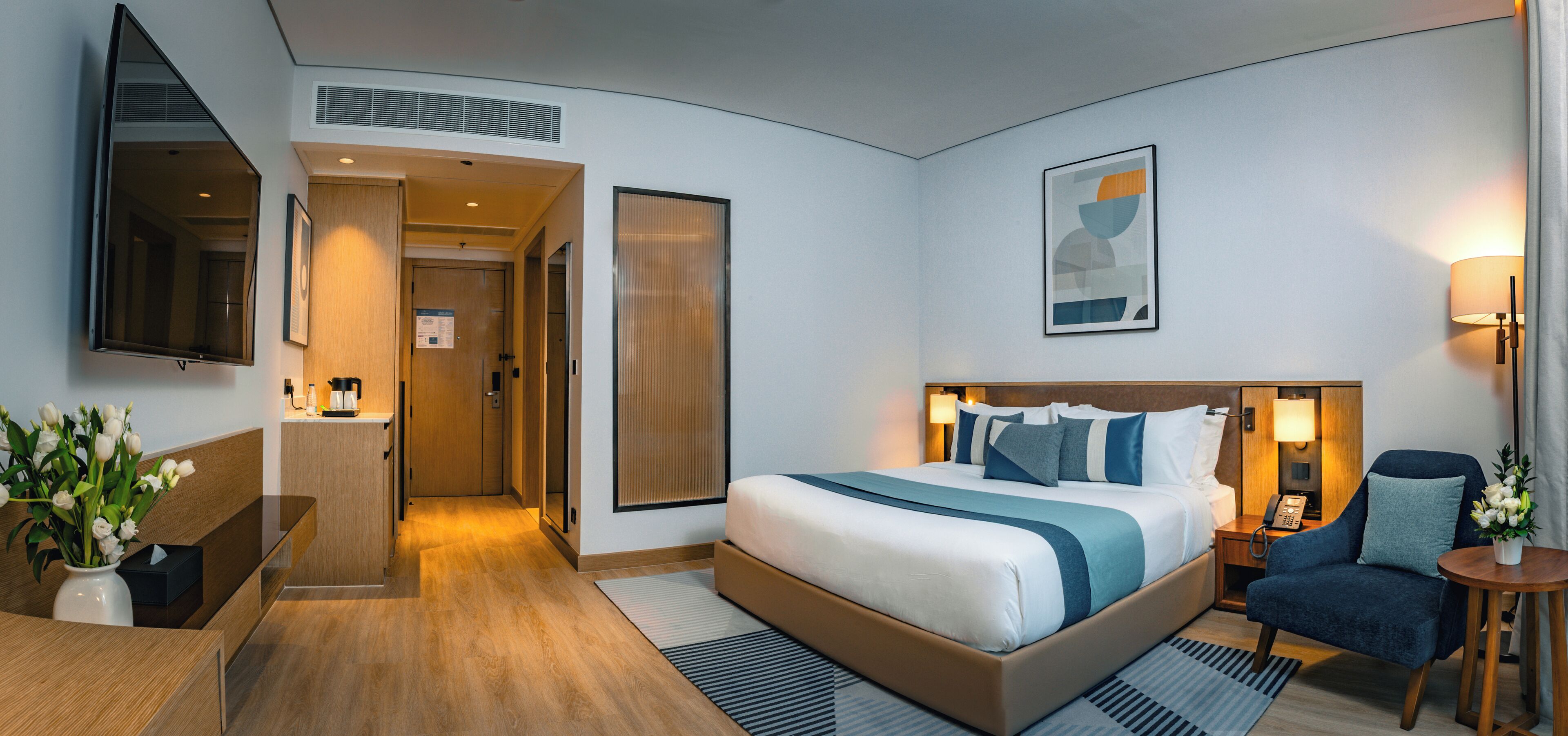 A modern hotel room with a large bed, bedside table, chair, and a desk with a vase of flowers.