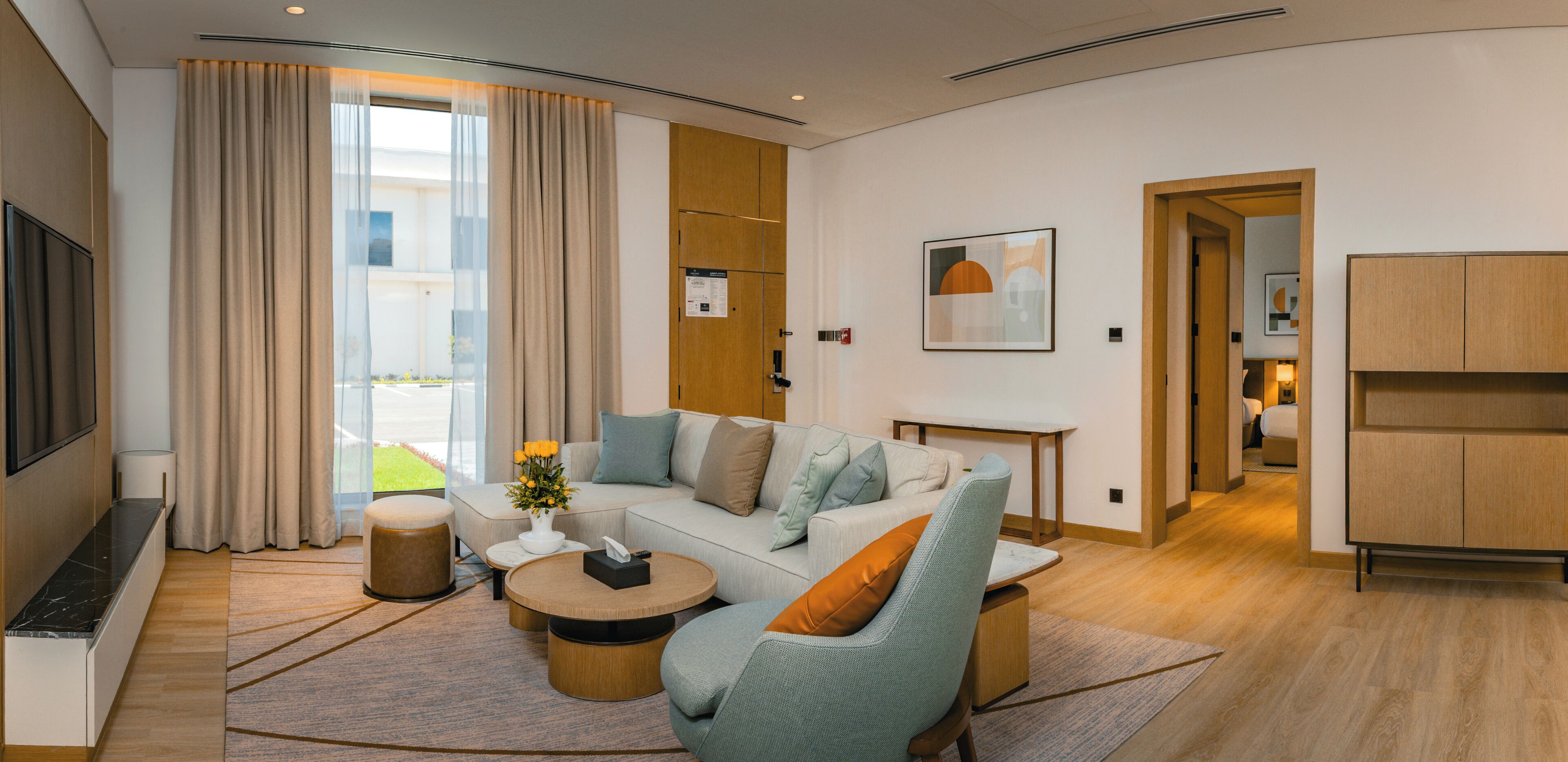A modern living room with a sofa, coffee table, and TV. There is a window with curtains, a door leading to another room, and a small table with a vase of flowers.