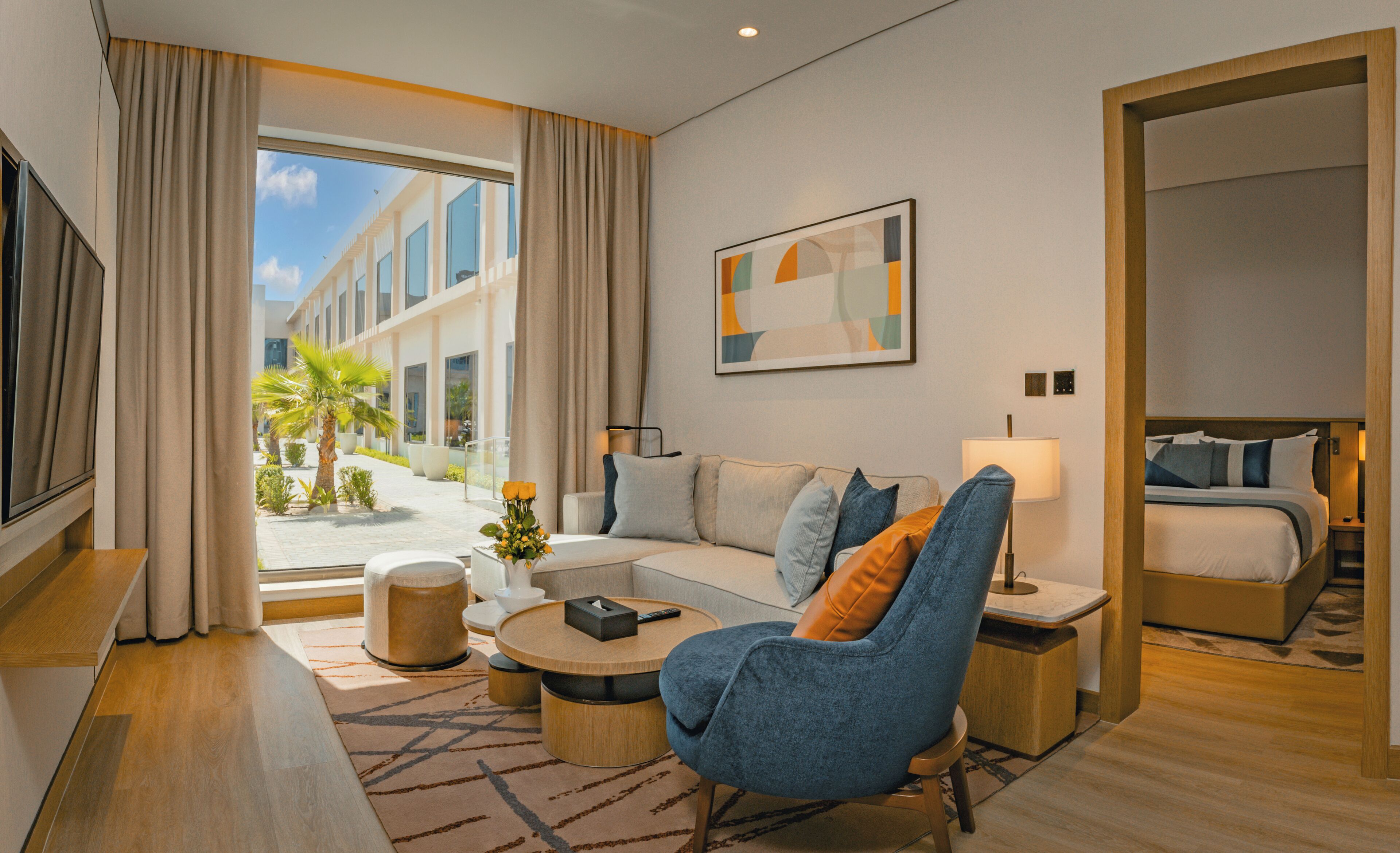 A modern hotel room with a sofa, armchair, and a bed. There is a window with curtains, a painting on the wall, and a small table with a vase of flowers.