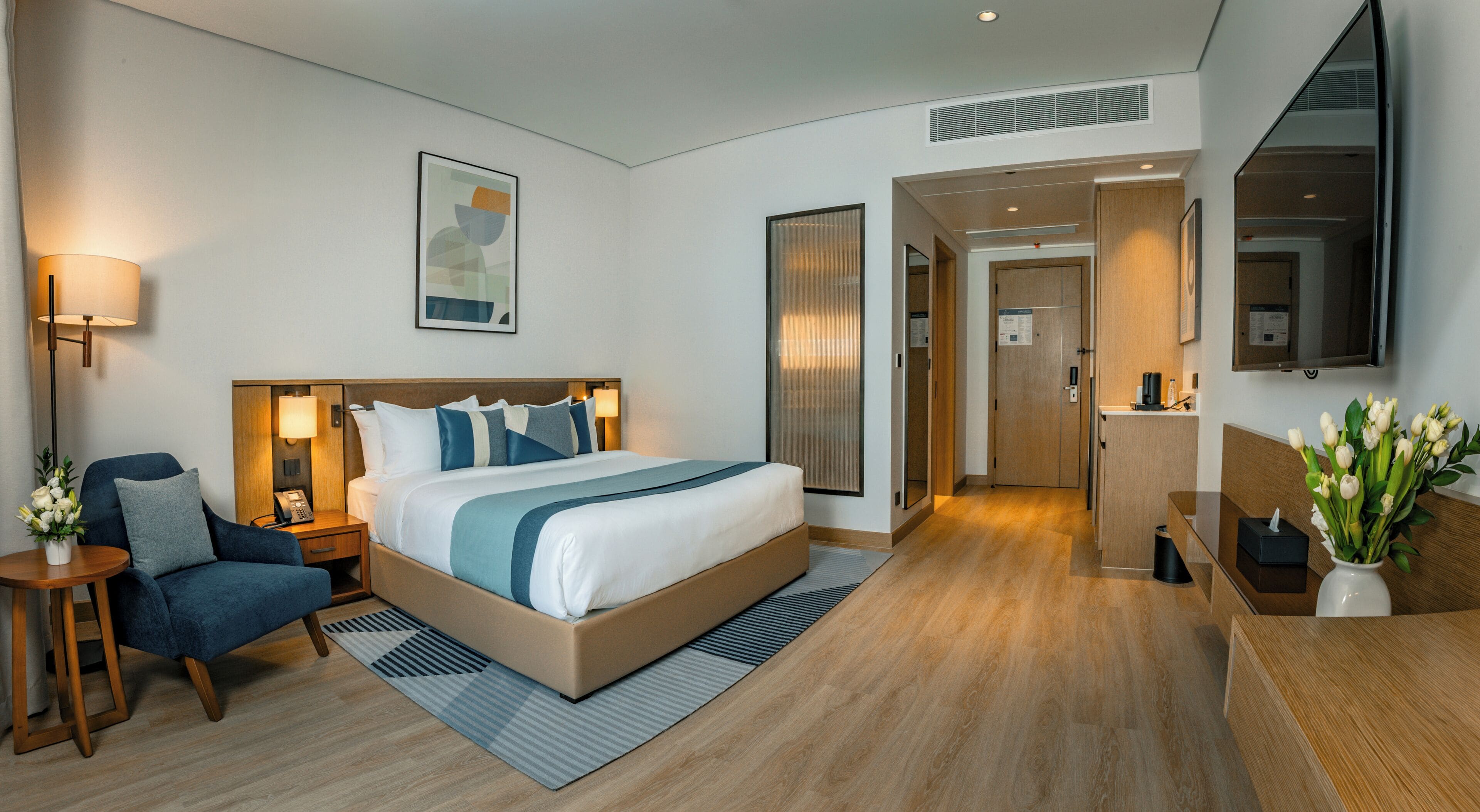 A modern hotel room with a large bed, a sitting area with a chair and a small table, a desk with a computer, a mirror, and a vase with flowers.
