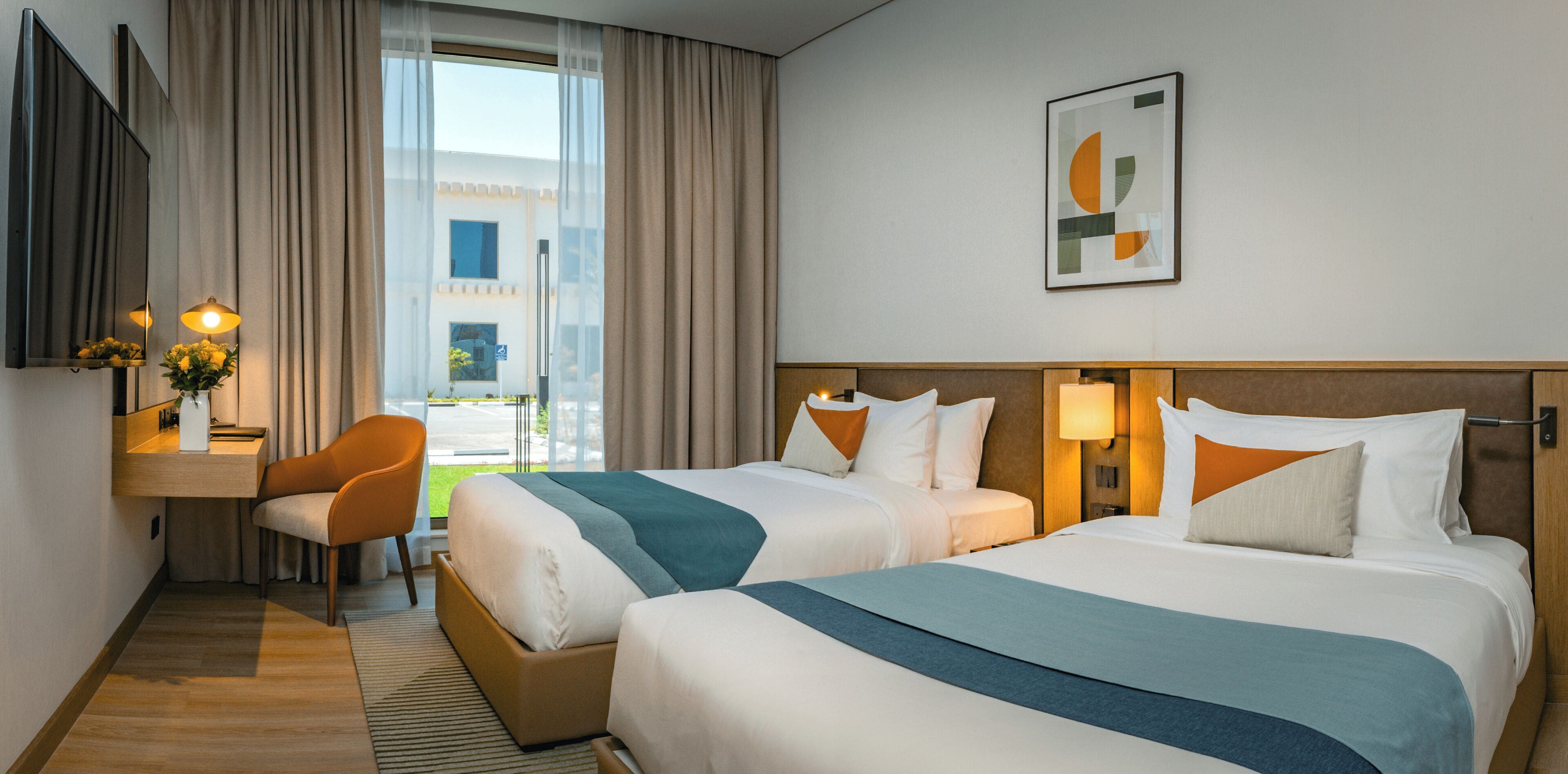 A hotel room with two beds, a desk, a chair, and a view of the outdoors.