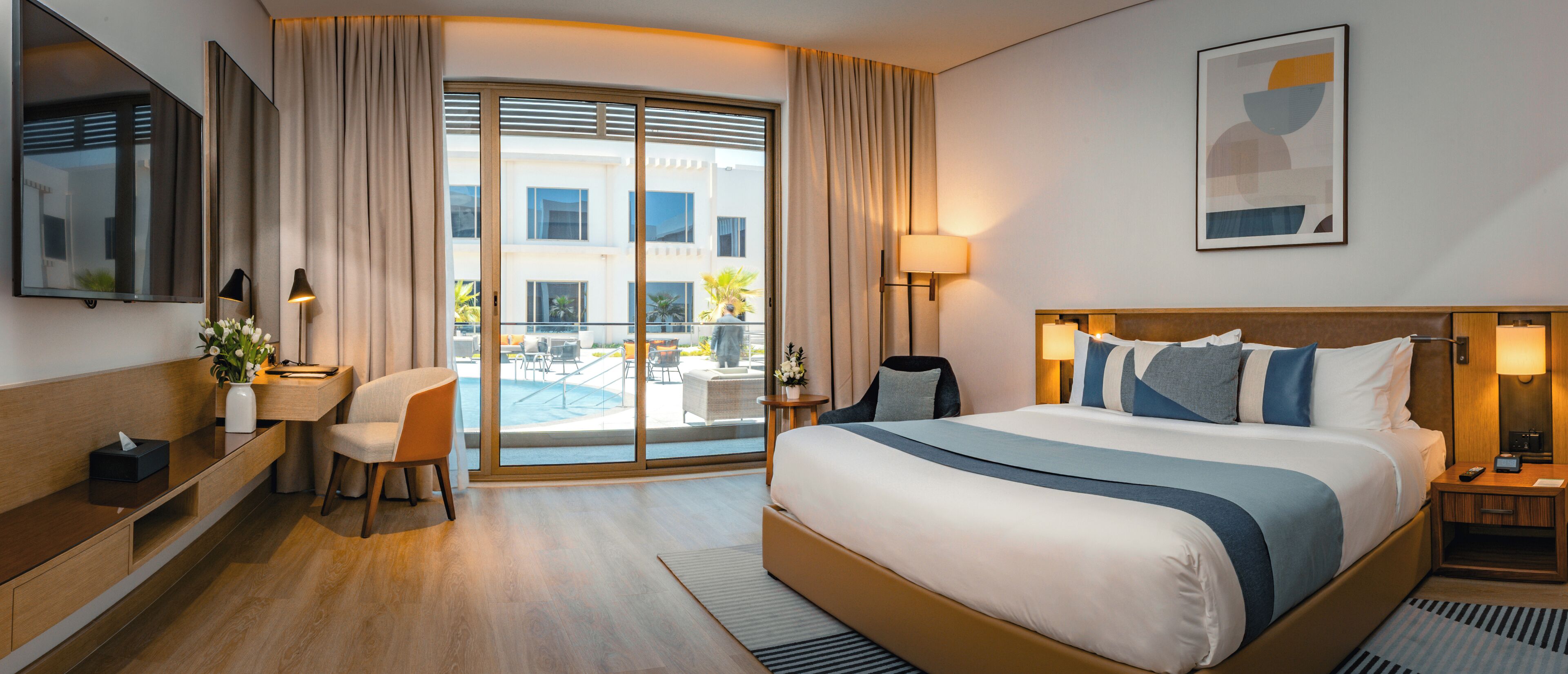 Une chambre d’hôtel avec un grand lit, un bureau avec une chaise, une petite table avec une lampe, et une vue sur la piscine et le bâtiment extérieur.