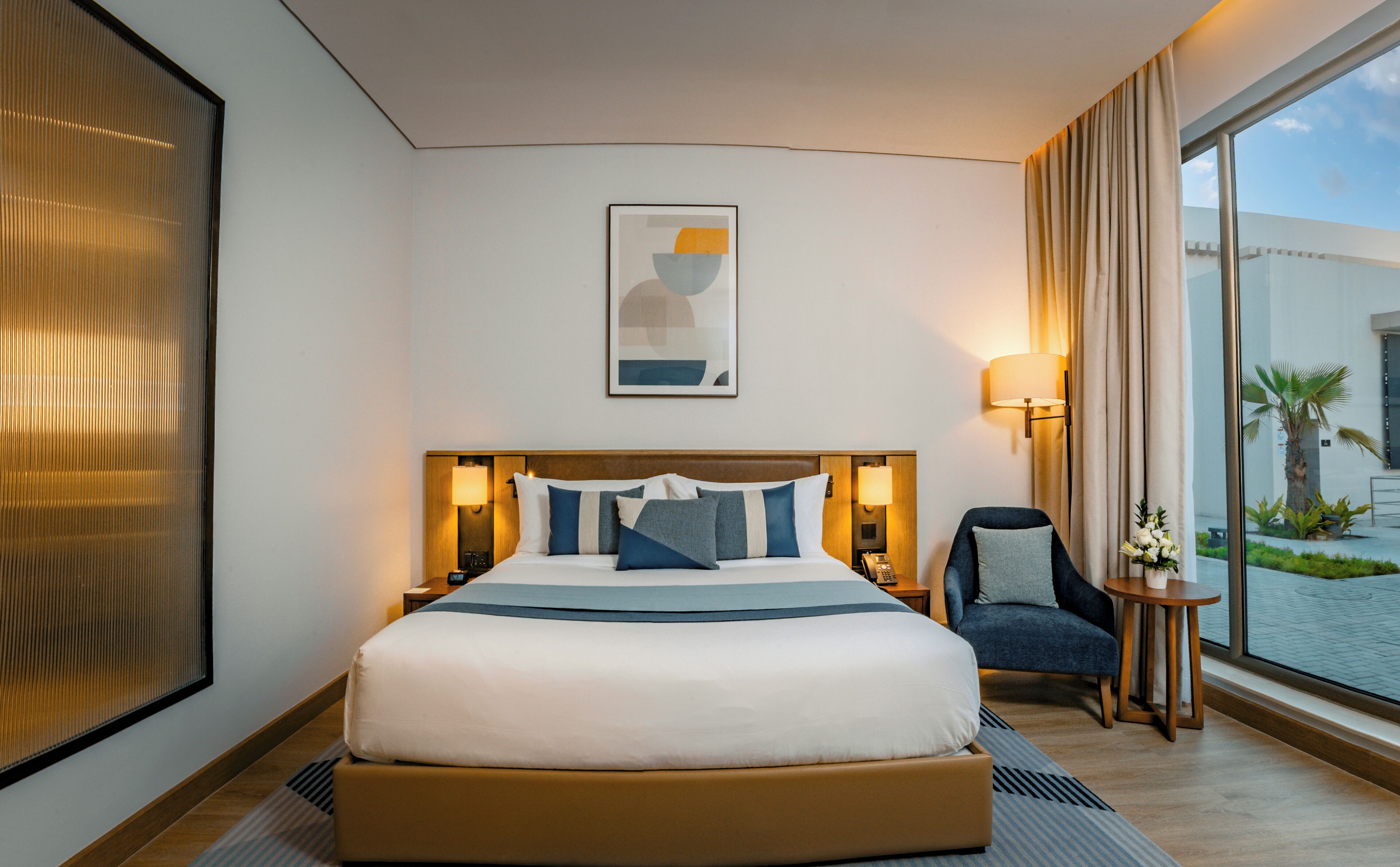 Habitación de hotel con una cama grande, una silla, una mesita de noche, una lámpara, un cuadro en la pared y vistas al exterior a través de una ventana.