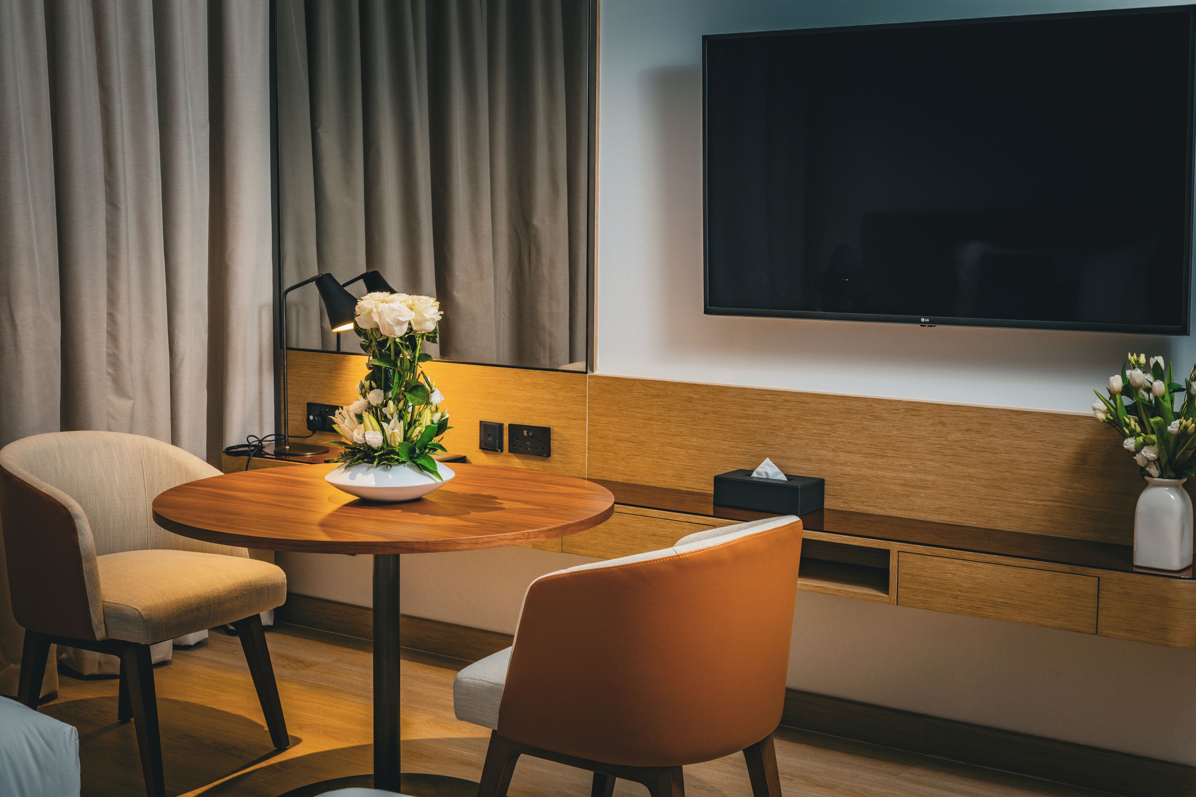 A modern hotel room with a round wooden table, two chairs, a flat-screen TV mounted on the wall, and a vase with white flowers on the sideboard.