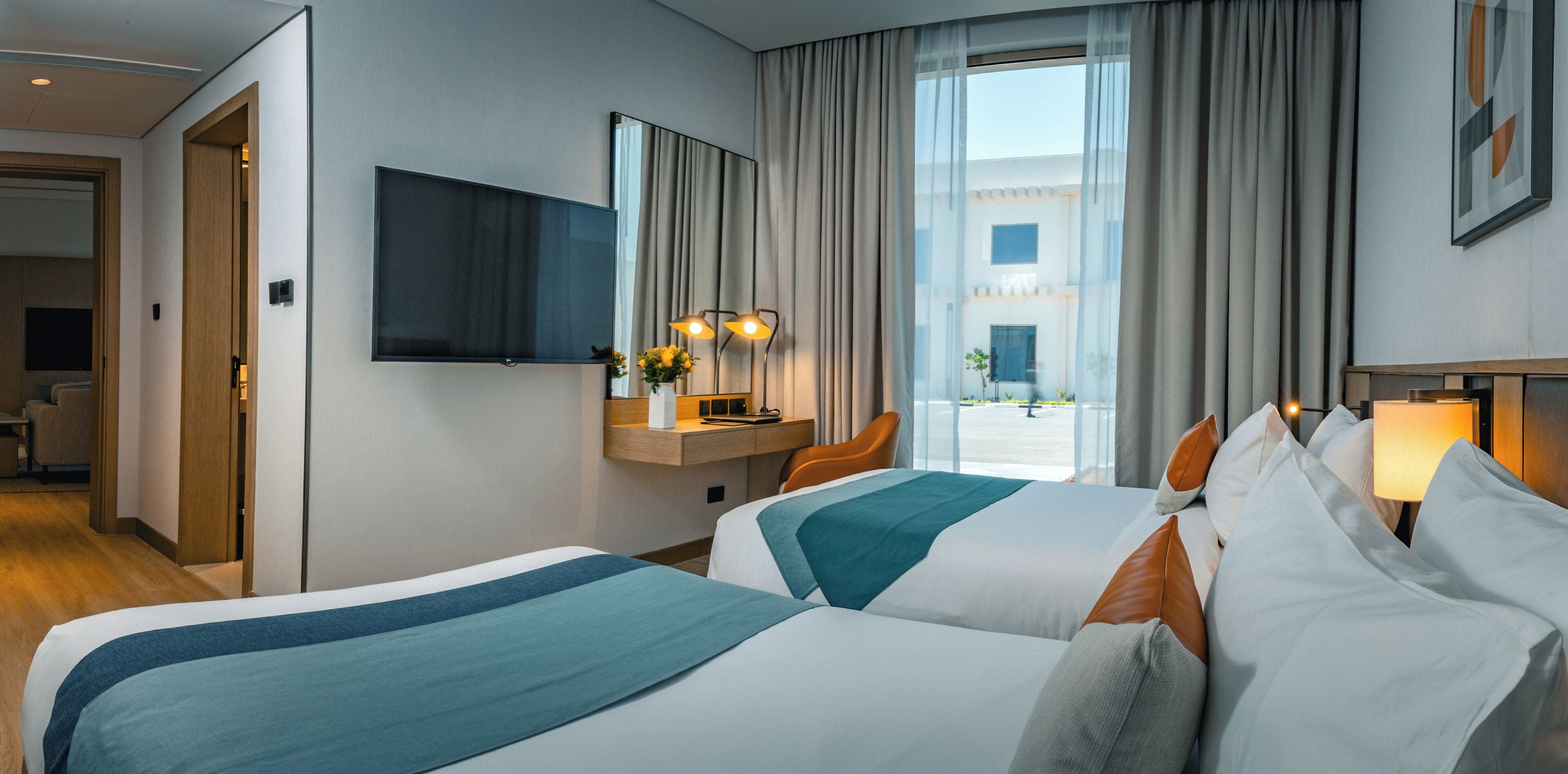 A hotel room with two beds, a desk with a chair, a TV, and a view of the outside.