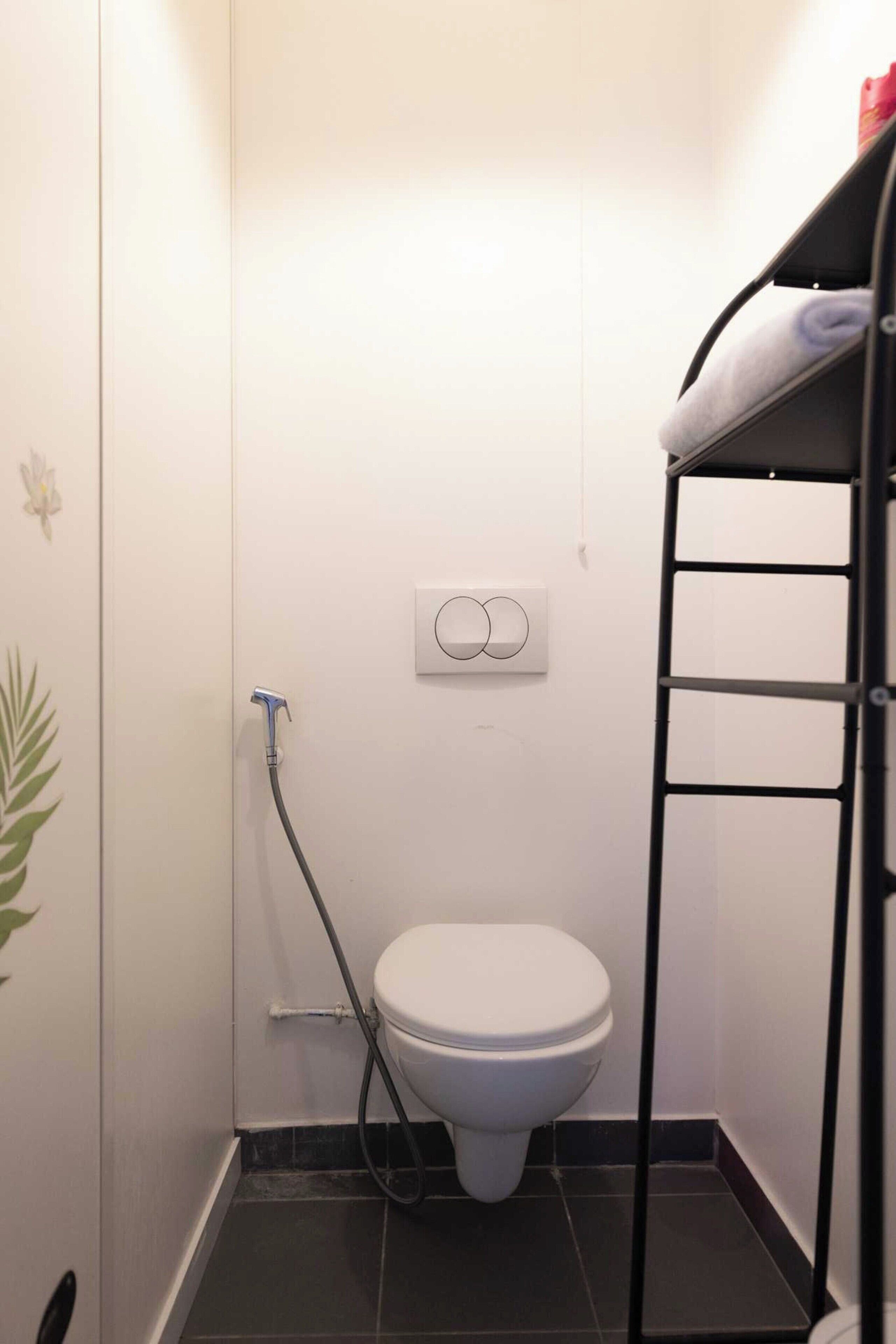 Une petite salle de bain avec des toilettes blanches, une étagère en métal noir et un tuyau de douche.
