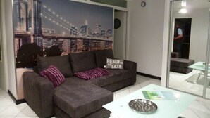 Ein Wohnzimmer mit grauem Sofa, einem Glastisch und einem Wandgemälde mit Stadtansicht.