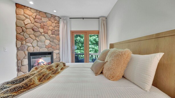 A bedroom with a stone wall, a fireplace, a bed with fluffy pillows, and a view of greenery through a glass door. - Walk to Holiday Valley, Family-sized House, Sleeps 9, Shuffleboard, 2 fireplaces (Ellicottville)