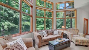 A spacious living room with large windows, a beige sofa, a patterned armchair, and a leather ottoman. - Walk to Holiday Valley, Family-sized House, Sleeps 9, Shuffleboard, 2 fireplaces (Ellicottville)