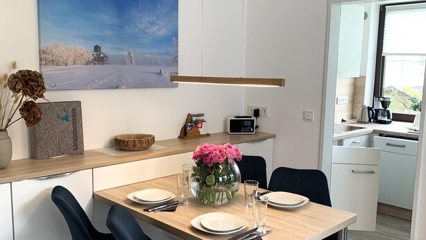 A modern kitchen with a wooden table set for two, two blue chairs, a vase with pink flowers, and a radio on the counter. - Winterberg gem With Pool, Sauna (Winterberg)