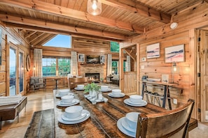 A wooden interior with a dining table set for four, a fireplace, and large windows.