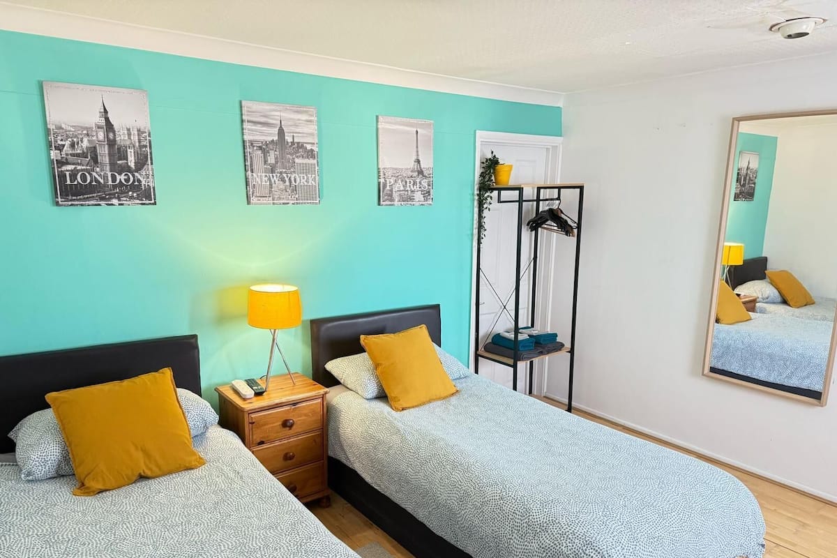 Quarto com duas camas, uma mesa de cabeceira com abajur, espelho e quadros com paisagens urbanas.