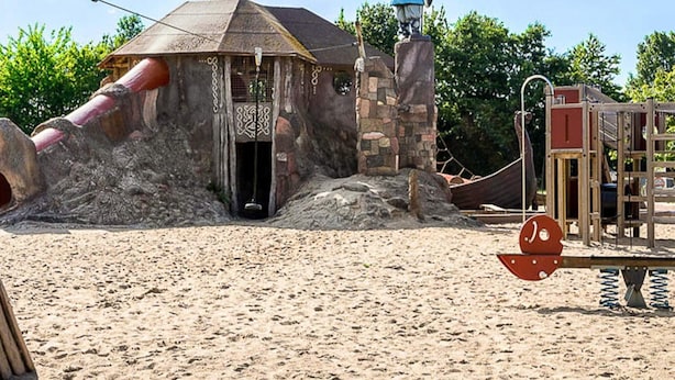Zona de jocs amb sorra, un petit edifici de pedra, una estàtua viking i un tobogan.
