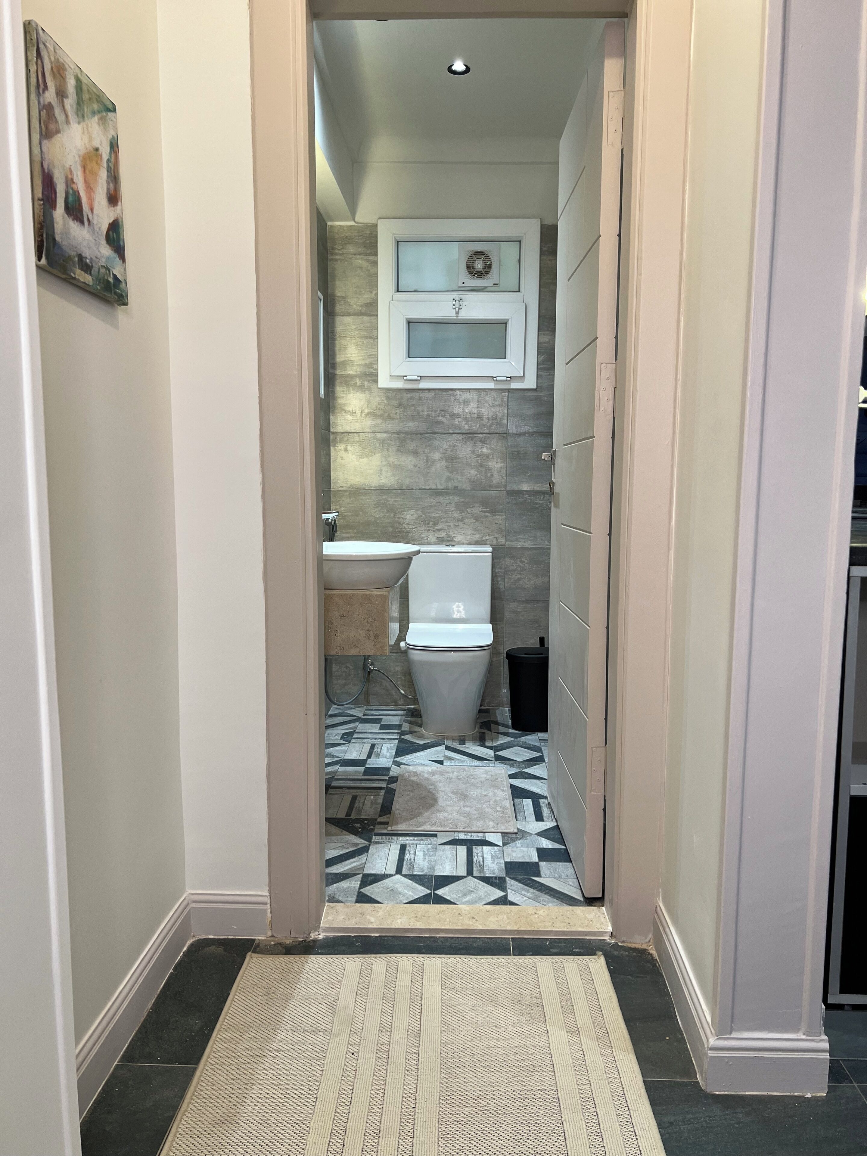 A narrow hallway leading to a bathroom with a tiled floor, a white toilet, and a mounted air conditioning unit.