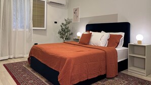 A bedroom with a bed, a nightstand, a bedside table, a potted plant, and a window with curtains. - 6th Of October Signature Stay (6th of October City)