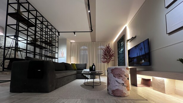 A modern living room with a sectional sofa, a round coffee table, a decorative pillar, a TV mounted on the wall, and a staircase with a metal railing. - 6th Of October Signature Stay (6th of October City)