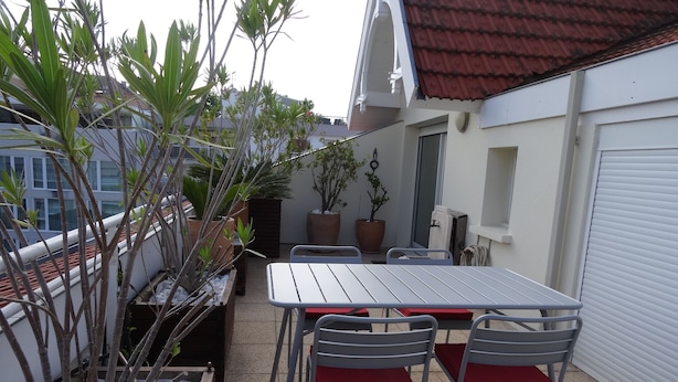 A patio with a table and chairs, surrounded by potted plants and a white building with a red roof.