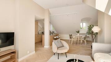 Un vaste espace de vie comprenant un téléviseur à écran plat, un canapé, une table à manger avec des chaises, et une cuisine à l’arrière-plan.