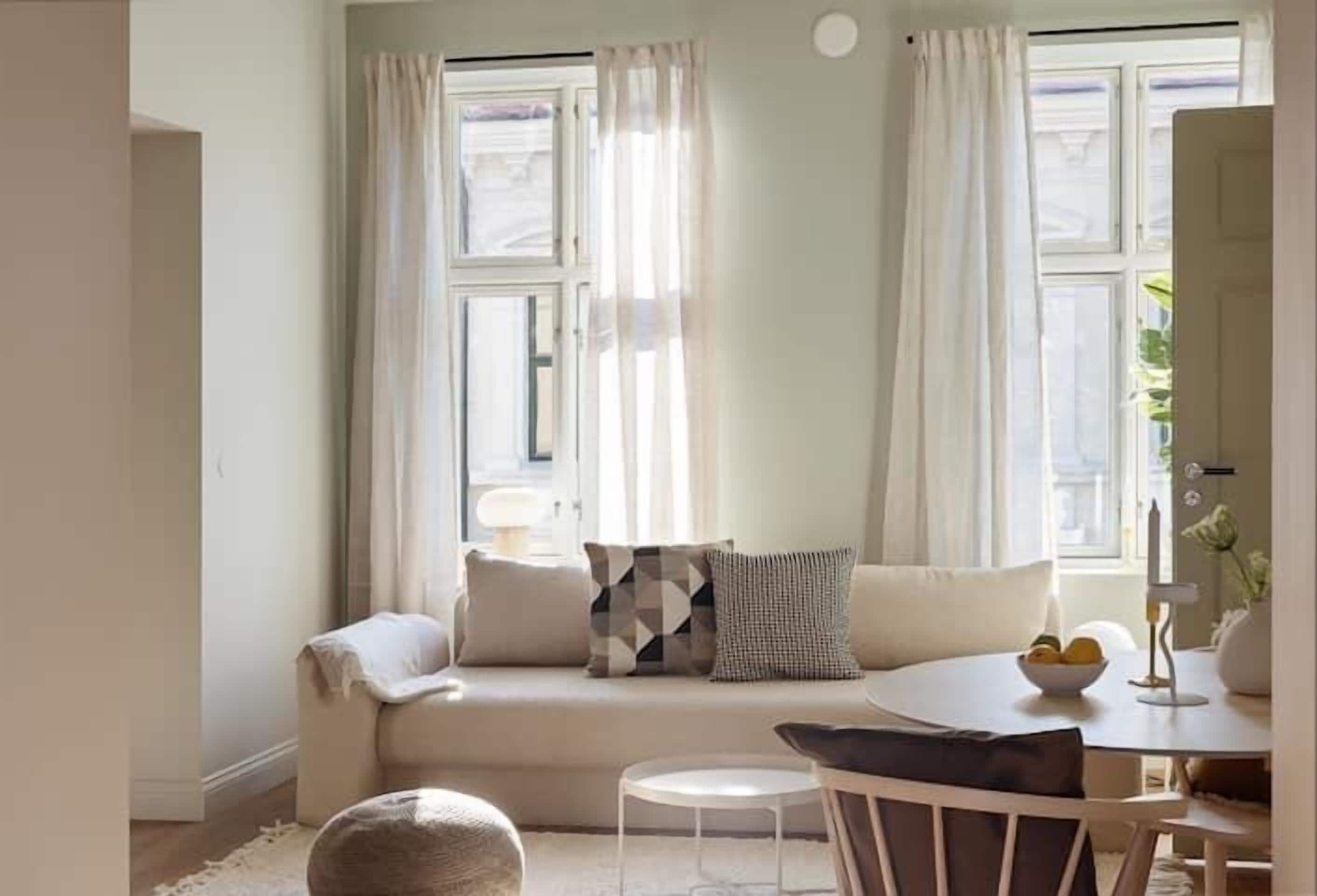 A cozy living room with a sofa, a round table, and a small white chair. There are two windows with curtains, a lamp on the table, and a bowl of fruit on the table.