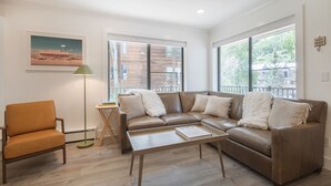 A modern living room with a brown leather sofa, a wooden coffee table, and a single orange chair. There are large windows providing natural light and a view of buildings outside. - Boomerang 202: New remodel! (Telluride)