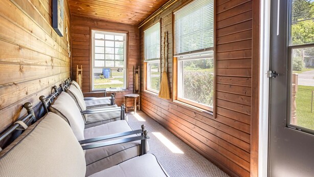 A cabin with wooden walls and ceiling, a row of beds with headboards, and large windows with blinds.