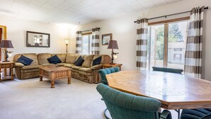 A living room with a sectional sofa, a wooden coffee table, a round dining table, and a teal armchair. There are two lamps on side tables, and the room has large windows with striped curtains.