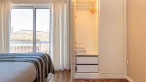 A bedroom with a bed, a window with curtains, a bathroom with a towel rack, and a view of buildings outside.
