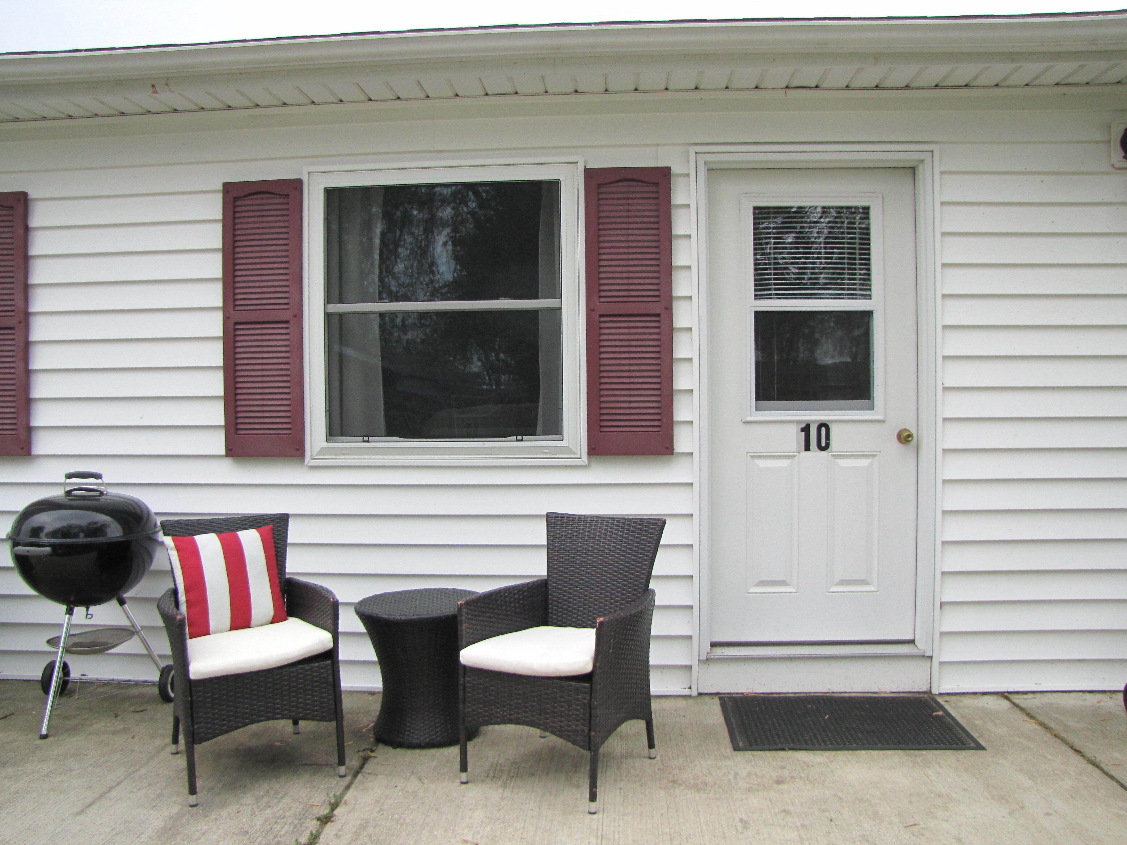 A house with a white exterior, a front door marked with the number 10, two wicker chairs with white cushions, and a barbecue grill.