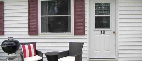 A house with a white exterior, a front door marked with the number 10, two wicker chairs with white cushions, and a barbecue grill.
