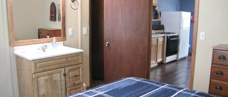 A bedroom with a bed, a wooden wardrobe, a bathroom with a mirror, and a kitchen visible through an open door.