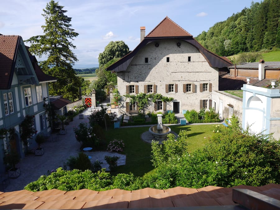 A traditional stone building with a courtyard, surrounded by lush greenery and a clear sky.