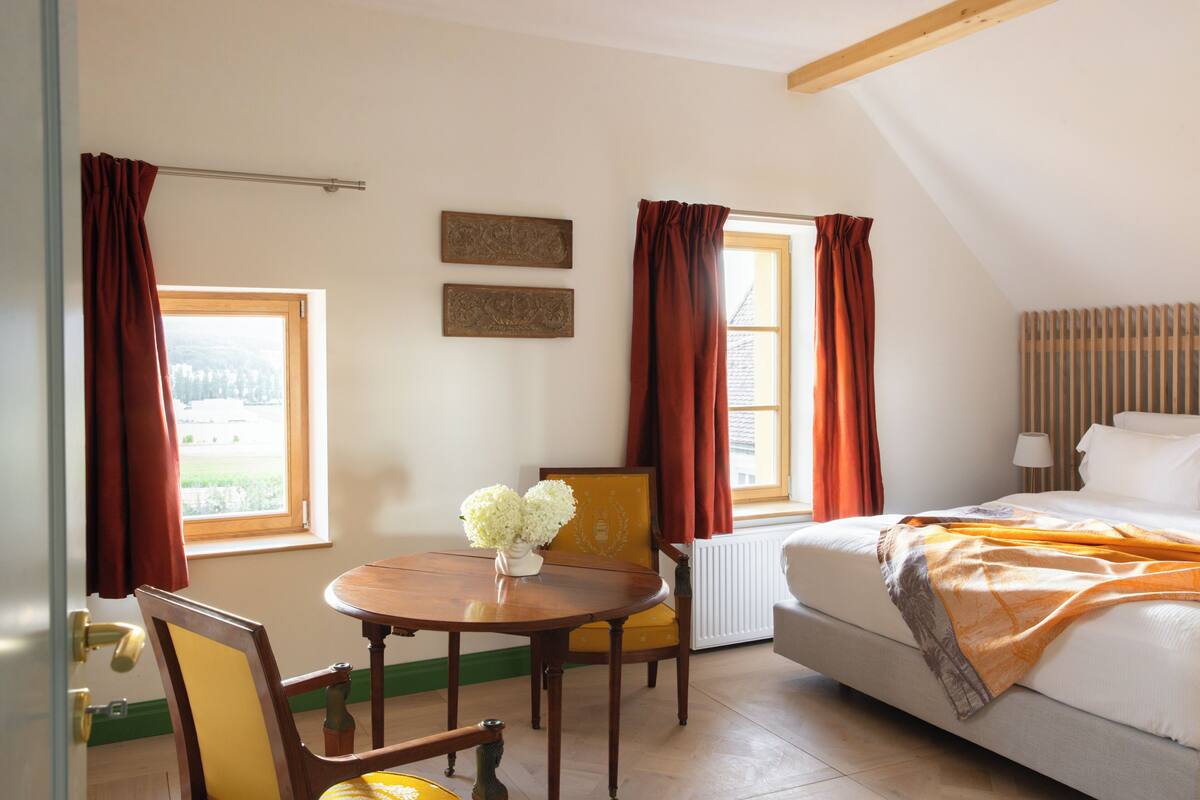 Ein Schlafzimmer mit einem Bett, einem Tisch mit Blumenvase und zwei mit Vorhängen versehenen Fenstern.