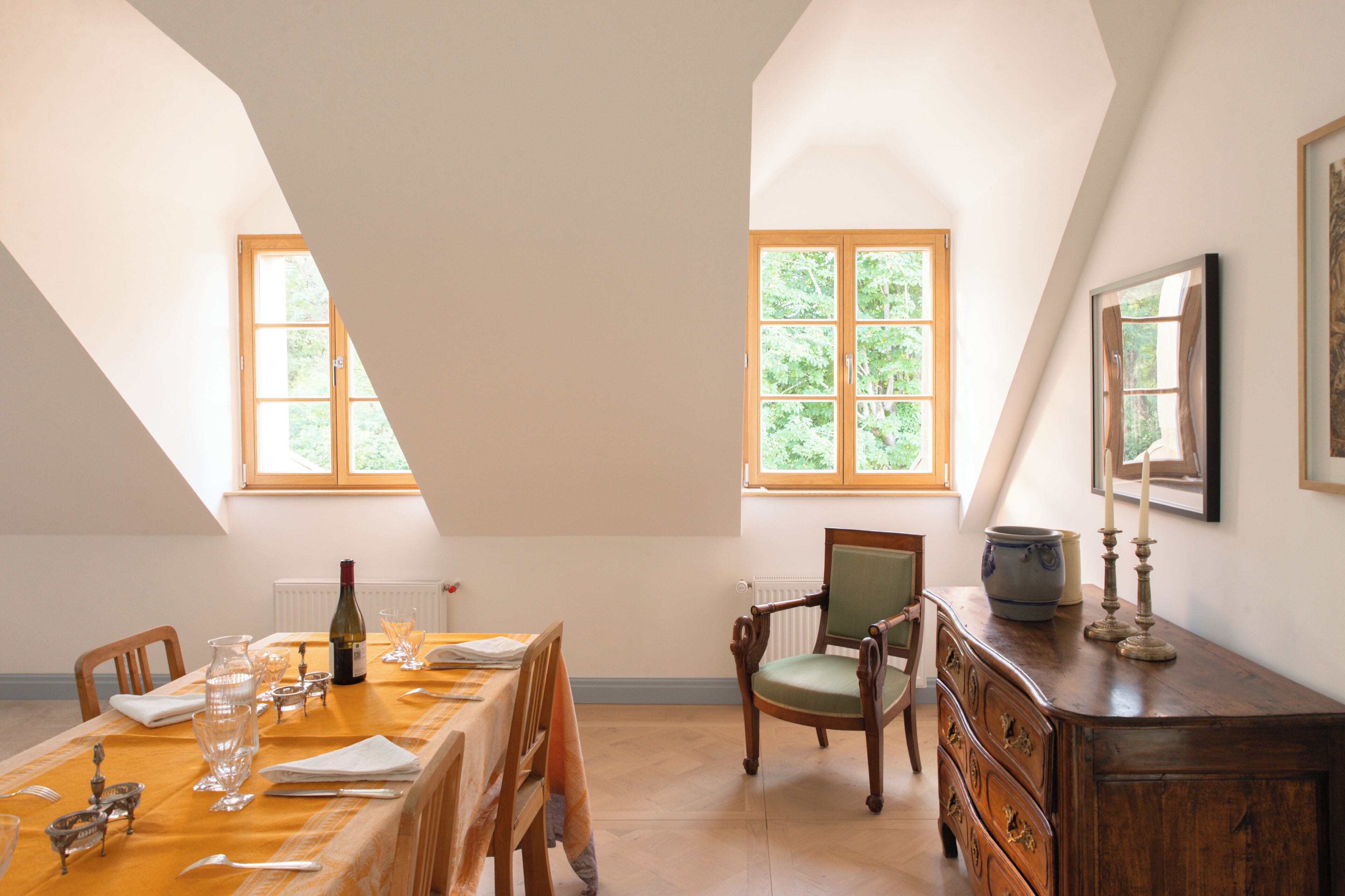 A dining room with a long table set for a meal, two windows, a wooden cabinet, and a chair.