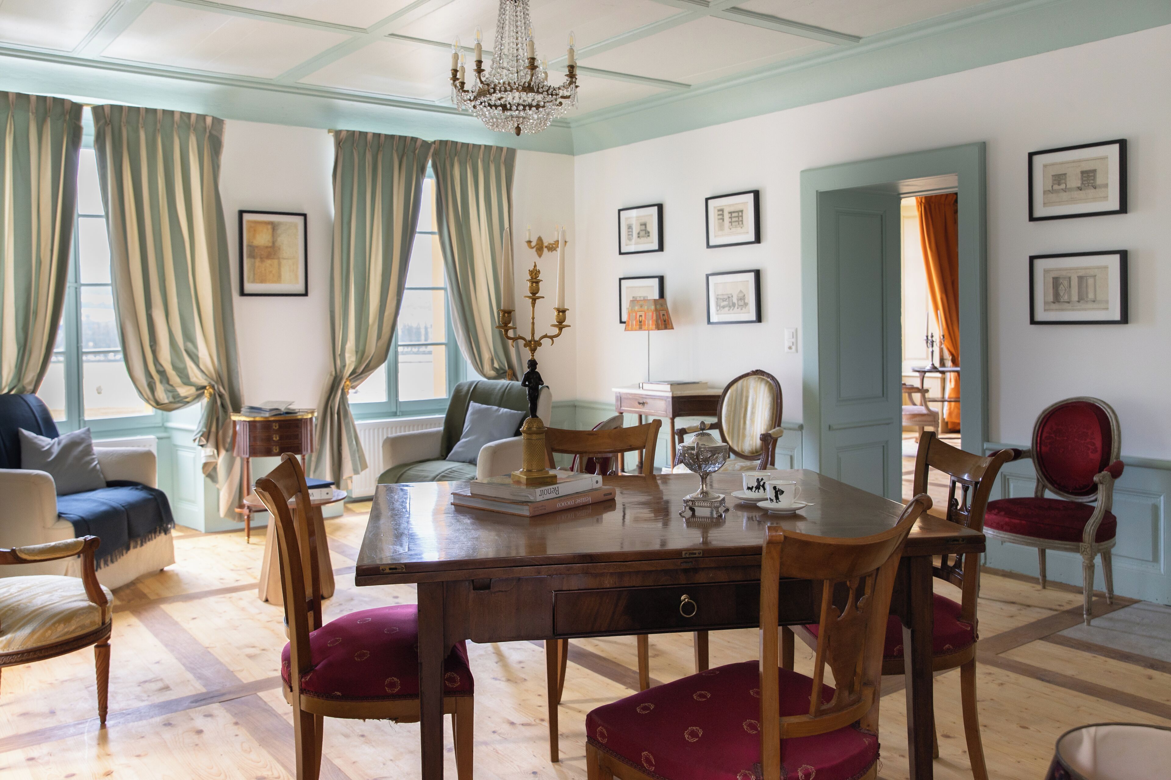Ein Esszimmer mit Holztisch, Stühlen und einer Chandelüster. Vorhänge, gerahmte Bilder an den Wänden und eine Tür zu einem anderen Raum sind ebenfalls zu sehen.