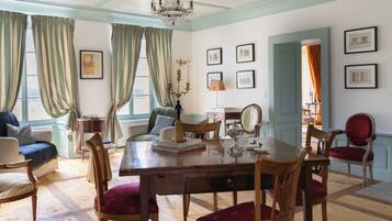 Ein Esszimmer mit Holztisch, Stühlen und einer Chandelüster. Vorhänge, gerahmte Bilder an den Wänden und eine Tür zu einem anderen Raum sind ebenfalls zu sehen.