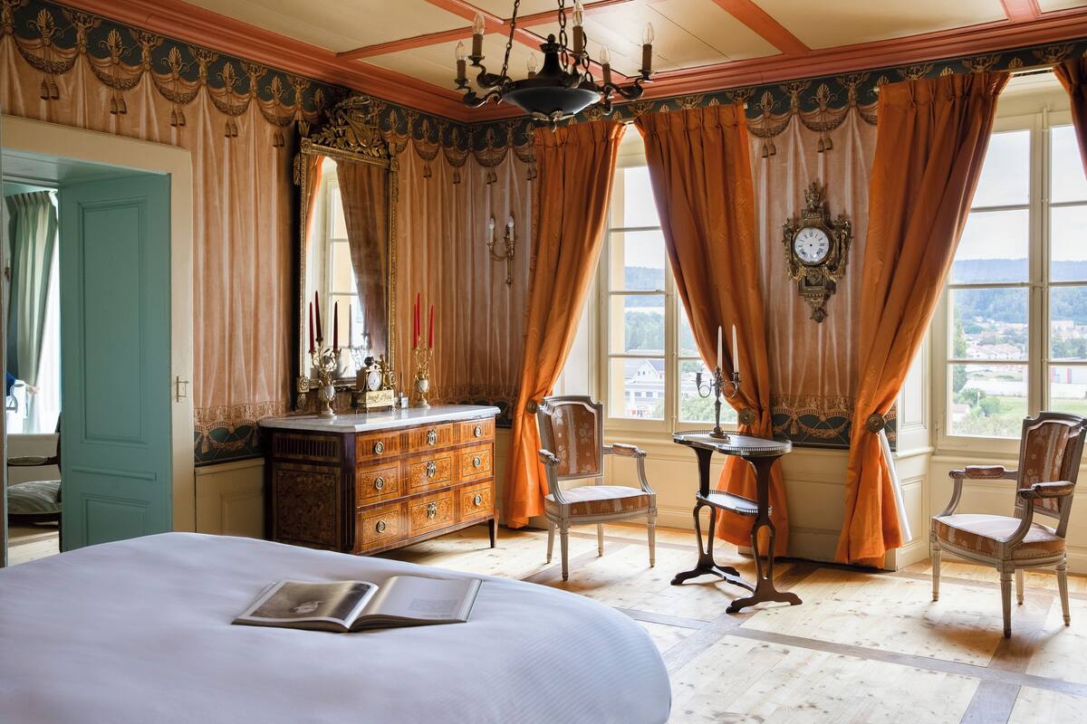 Ein geräumiges Schlafzimmer mit einem großen Bett, einem Schminktisch, zwei Stühlen, einem Tisch und Blick ins Freie durch große Fenster.