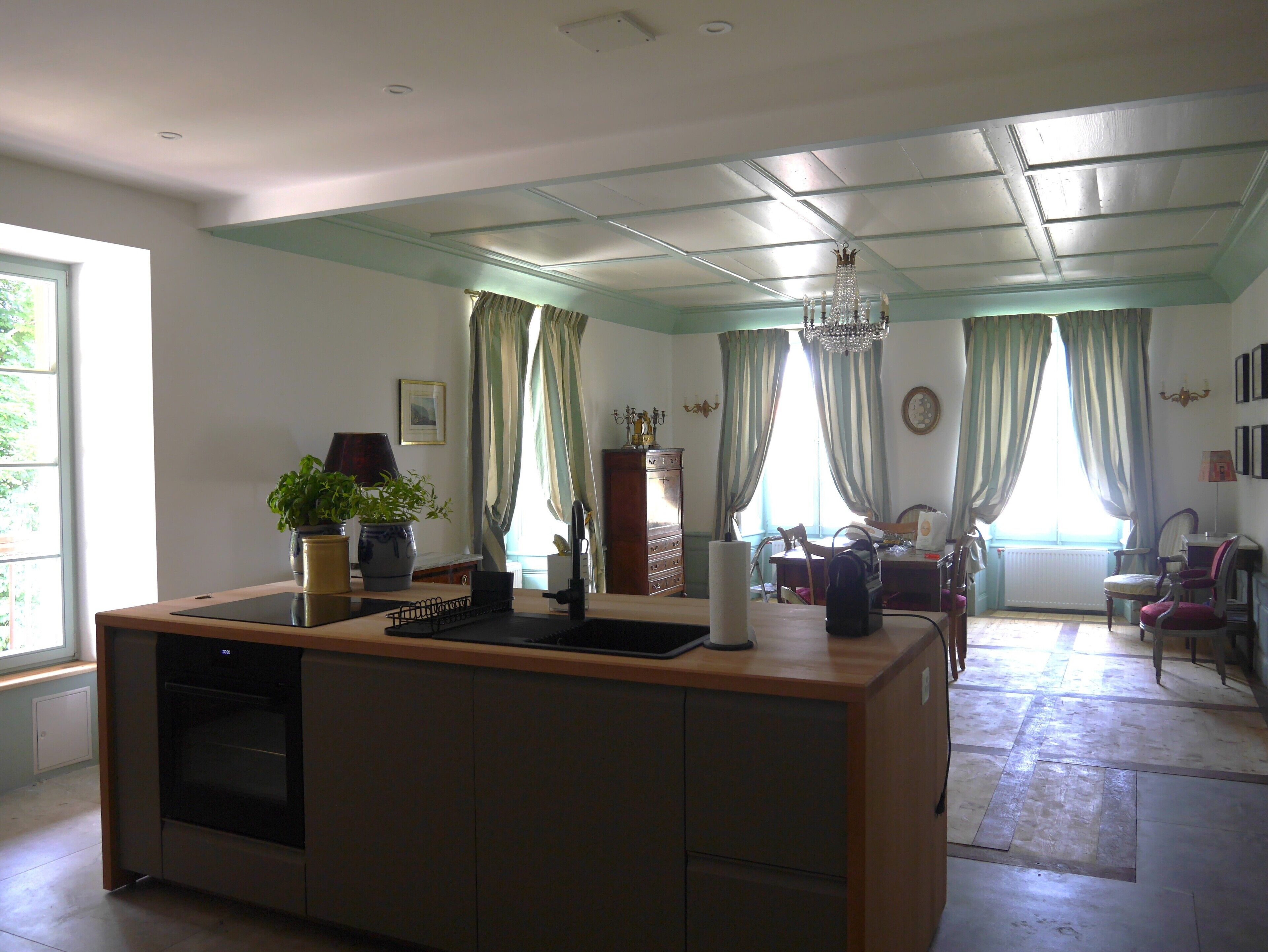 A modern kitchen with a built-in oven, a dining area with a chandelier, and a view of the outdoors through large windows with curtains.