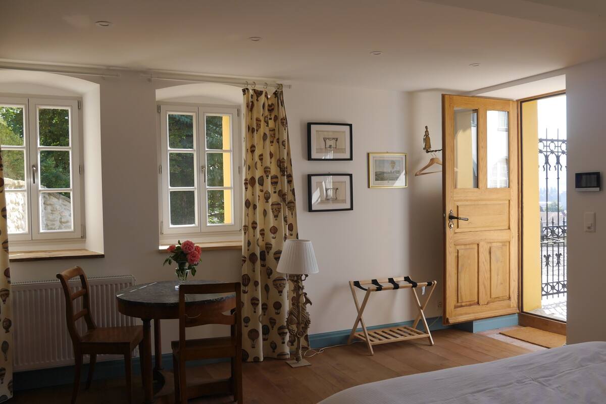 Ein Zimmer mit einem Bett, einer Holztür, einem Fenster mit Vorhängen, einem Tisch mit Stühlen und gerahmten Bildern an der Wand.