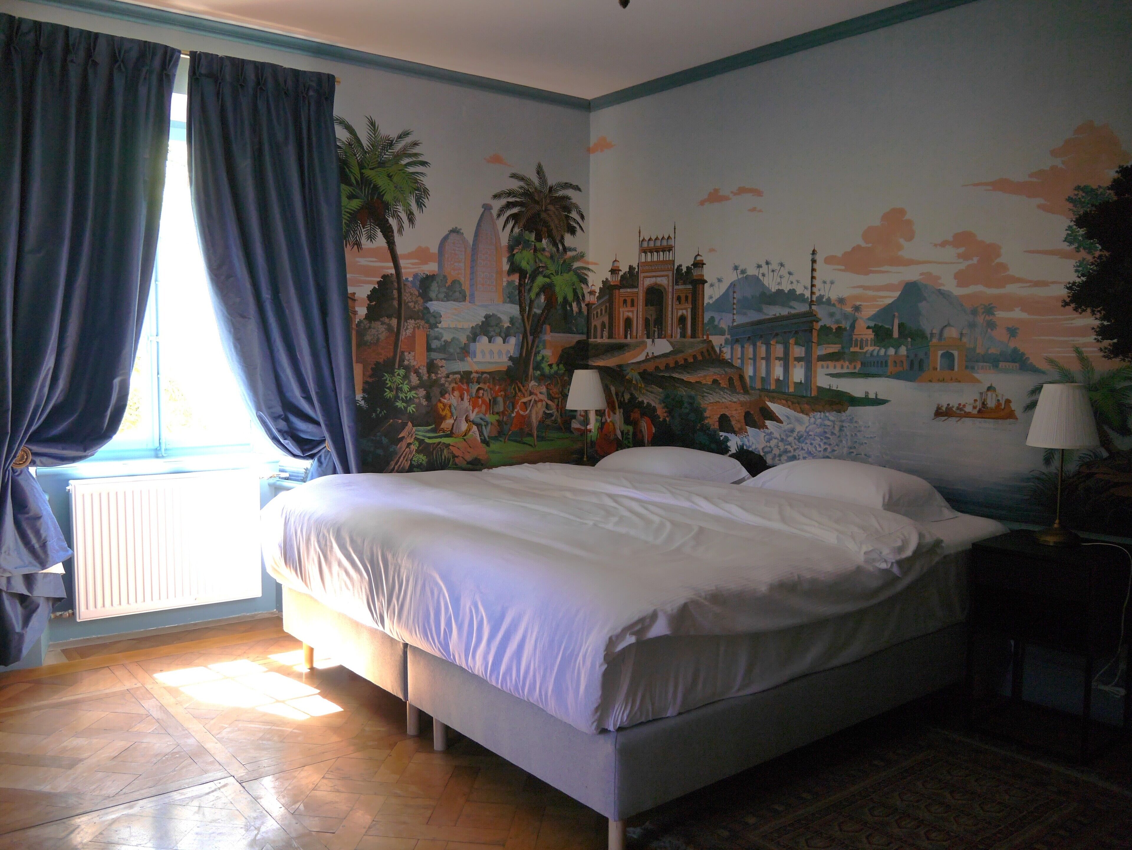 Ein Hotelzimmer mit einem großen Bett, einem Wandgemälde, das eine historische Szene zeigt, einem Fenster mit Vorhängen und einem Heizkörper.
