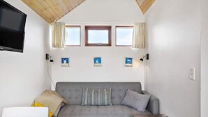 A cozy room with a grey sofa, a small wooden table, and a TV mounted on the wall. There are three windows with curtains, and a few small pictures on the wall. - 5 Person Holiday Park Home in Krusa (Krusaa)