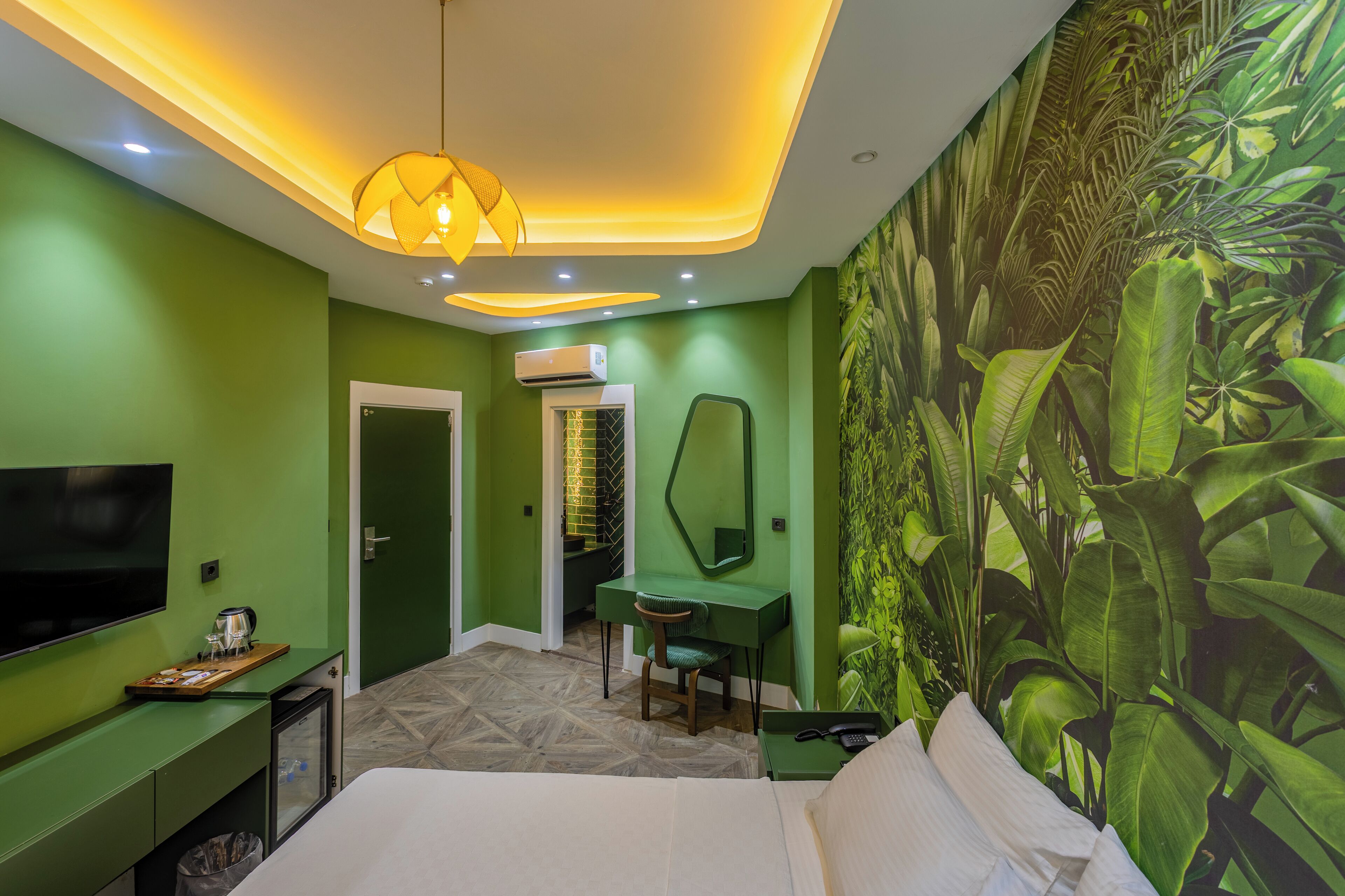 A hotel room with a bed, a desk, a chair, a TV, and a wall with a green leafy mural.