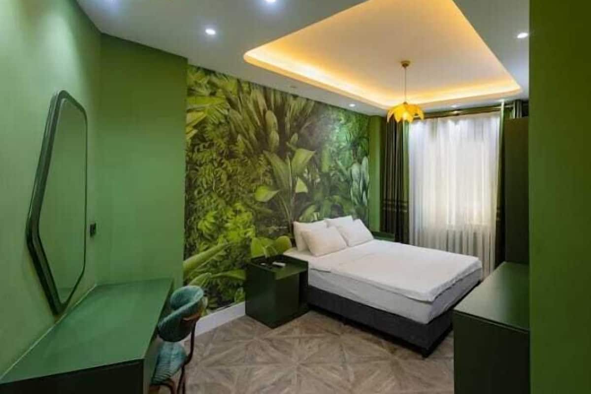 A bedroom with a bed, a nightstand, a green wall with a jungle mural, and a ceiling light fixture.