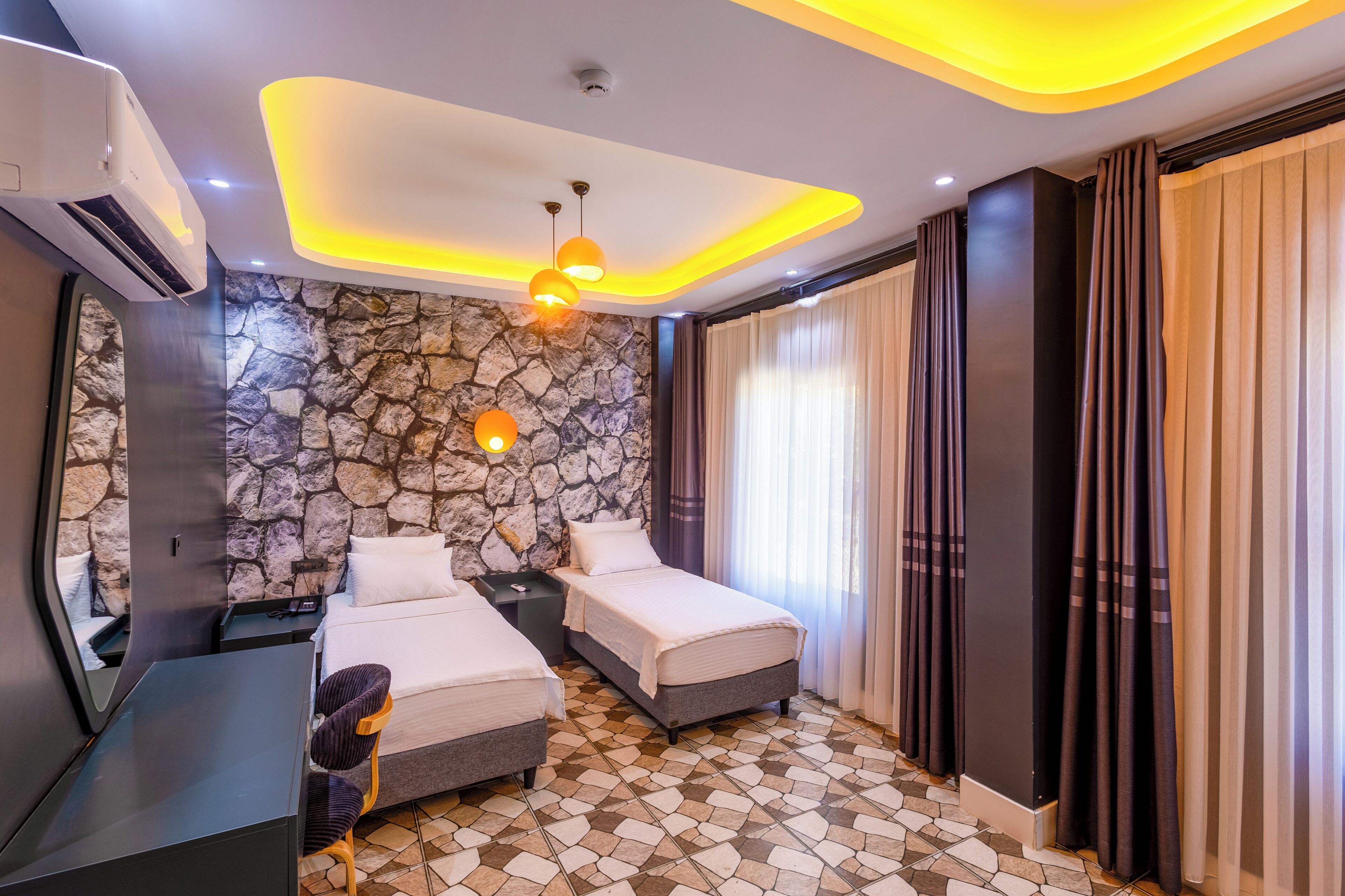 Una habitación de hotel moderna con dos camas, una pared decorativa de piedra, piso de baldosas e iluminación empotrada en el techo.