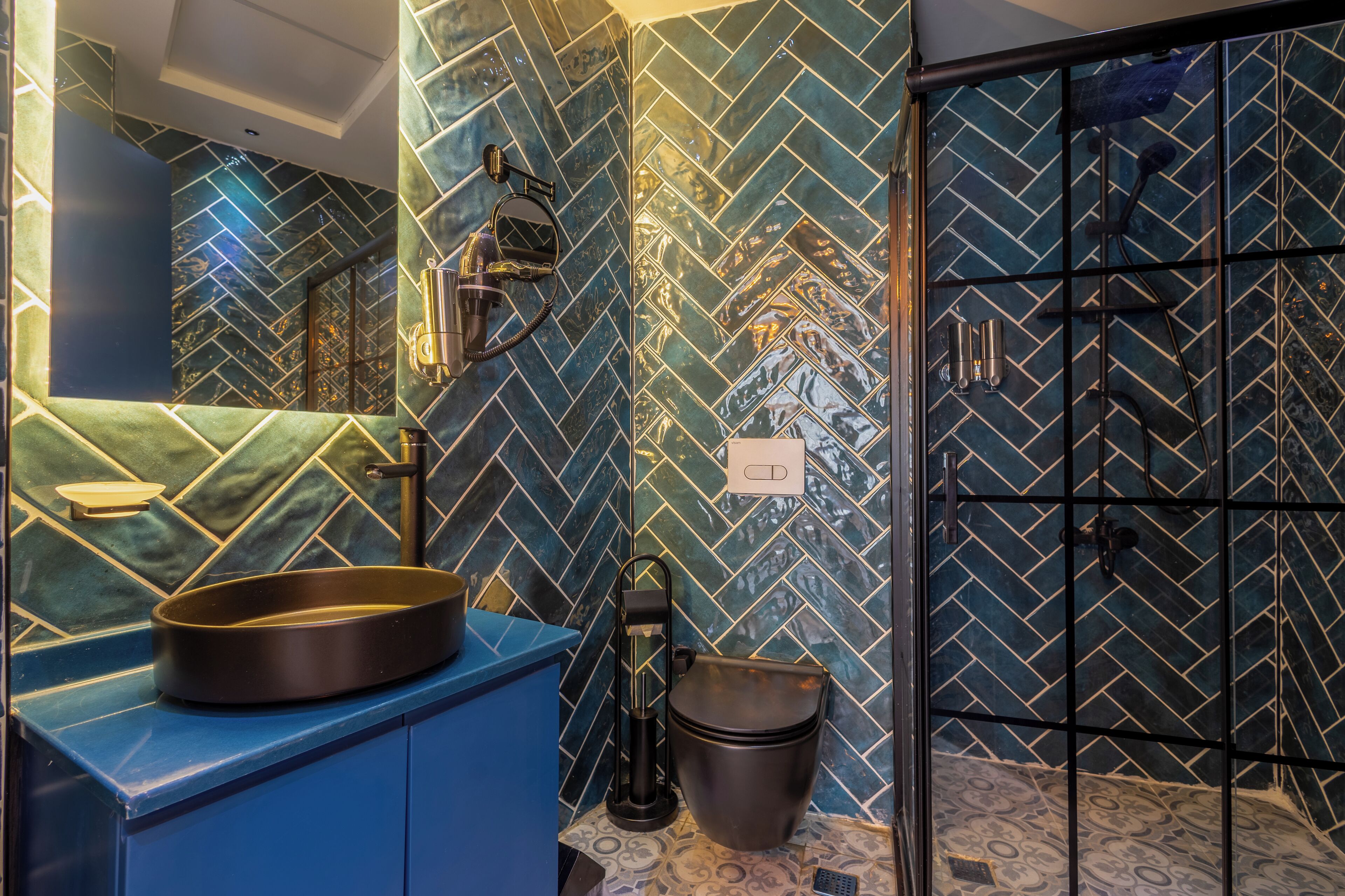 Un baño con un patrón de baldosas de mosaico azul y verde tipo espiga, un lavabo negro y un box de ducha con puerta de rejilla negra.