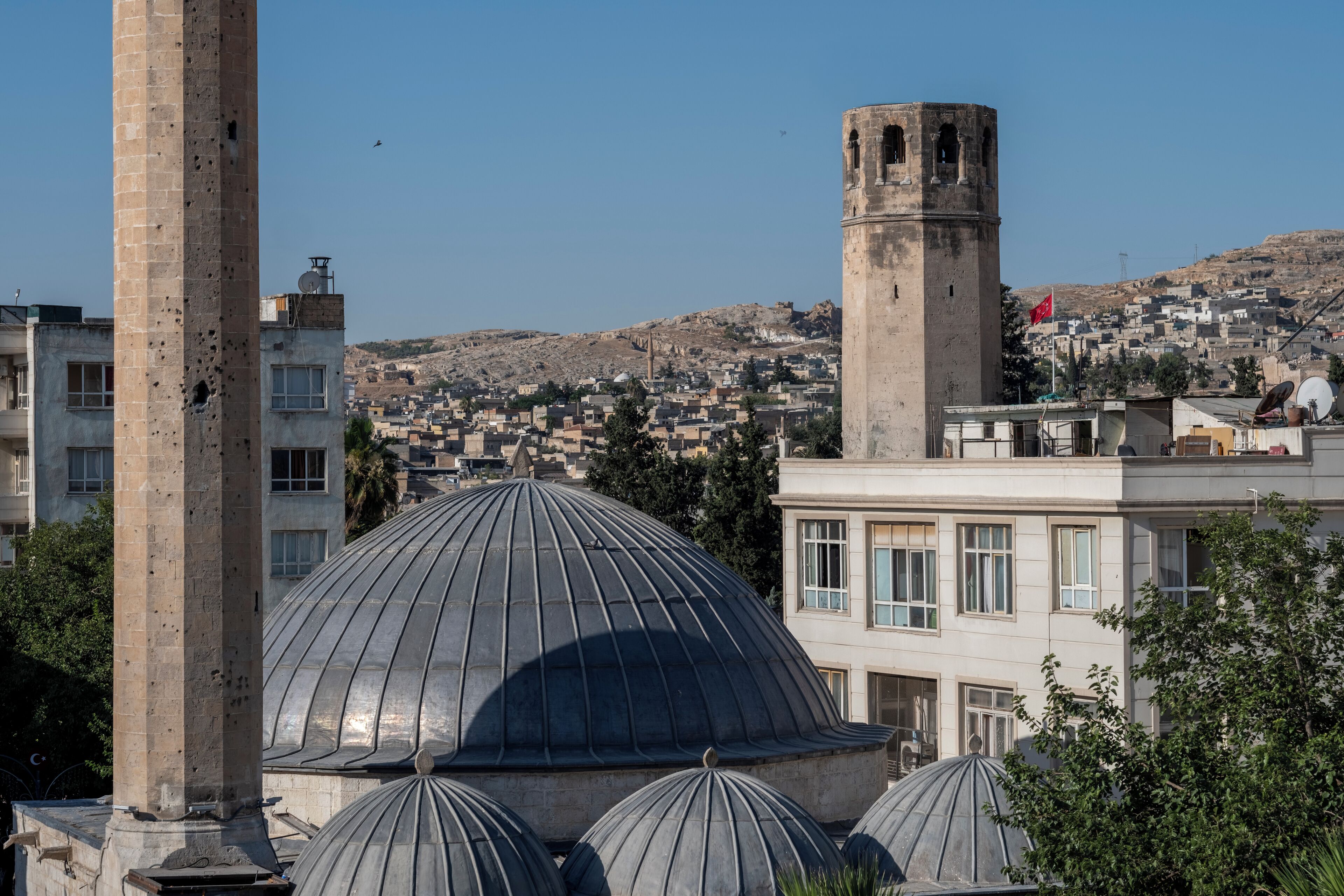 A mosque with large domes and a minaret, surrounded by residential buildings and a hilly landscape.