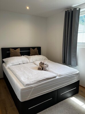 A neatly made bed with white linens and rolled towels, a black headboard, and a window with grey curtains.